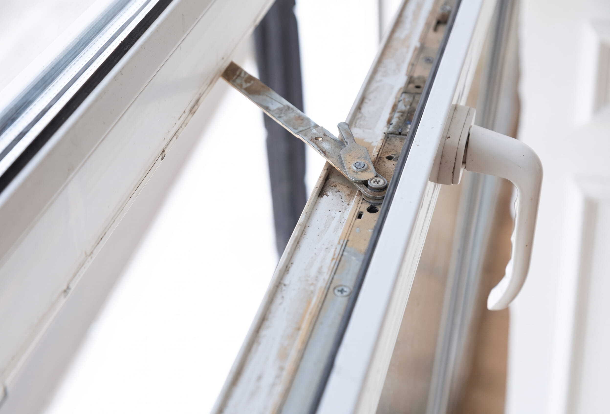 An open, weathered white uPVC window on a slight angle, showing the inner hardware, stay arm, and handle. Natural light streams through, highlighting some dust and wear on the tracks, illustrating the need for maintenance and repairs. 